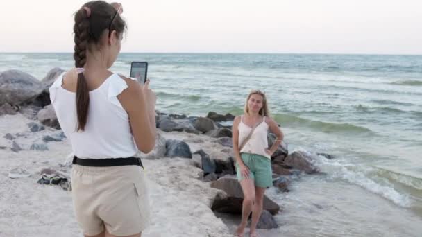 太いサングラスと携帯電話を手にした少女が 海岸の石砂のビーチで石の上に座っている彼女の豪華な母親の写真を撮っている Uhdスローモビデオ — ストック動画