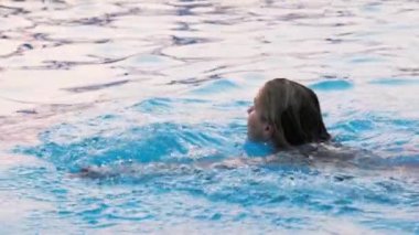 Beautiful young tanned woman swims in a deep blue pool with clear transparent cool water against the backdrop of a bright summer sunny sunset on a warm calm vacation. HD slow-mo video