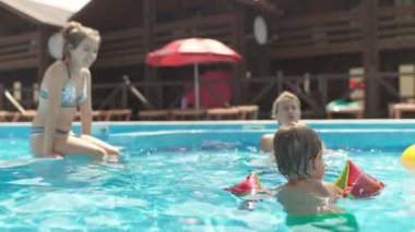 Parlak şişme kolluklu neşeli çocuk, sığ mavi bir havuzda temiz şeffaf bir suyla, ilgili bir anne ve ablasıyla sıcak bir yaz tatilinde yüzüyor. HD yavaş çekim videosu