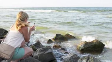 Beautiful happy young woman in mild light clothes with a careful braid, takes pictures of the landscape of the sea horizon on a modern phone while sitting on wet stones. HD slow-mo video