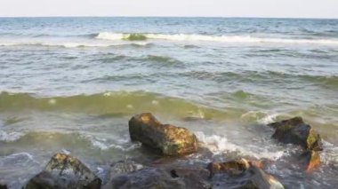 Many large and small stones lie on the wide large stone shore of the stormy blue Sea of Azov, and the dark foamy majestic waves break against the boulders. HD slow-mo video