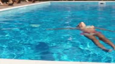 Açık pembe bir mayo giymiş, çocuklar için mor gözlüklü, ince bir kız, temiz şeffaf suyun üzerinde sırtüstü uzanıyor ve ılık güneşli ve yaz tatillerinin tadını çıkarıyor.