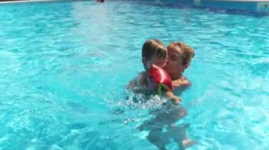 Happy caring kind mother in a bright bikini, plays and swims with a funny baby in oversleeves and his older sister in a deep blue pool with clear transparent water. HD slow-mo video