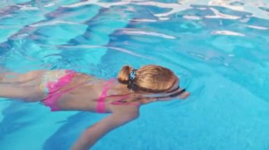 Yüzmek için şeffaf siyah gözlüklü, parlak pembe bir mayo giyen küçük bir kız gözlüklerini düzeltiyor ve berrak bir su şemsiyesiyle birlikte derin mavi bir havuza dalıyor. HD yavaş çekim videosu