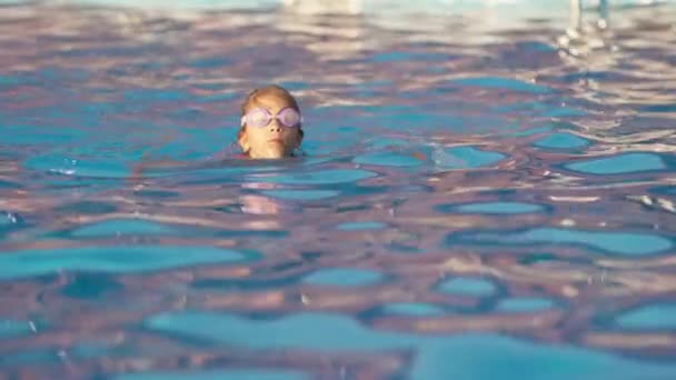 一个身穿亮晶晶粉色泳衣的小女孩带着透明的黑色护目镜游泳 把护目镜伸直 然后沉入一个深蓝色的池子里 里面有一串串清澈的水 Uhd慢速视频 — 图库视频影像