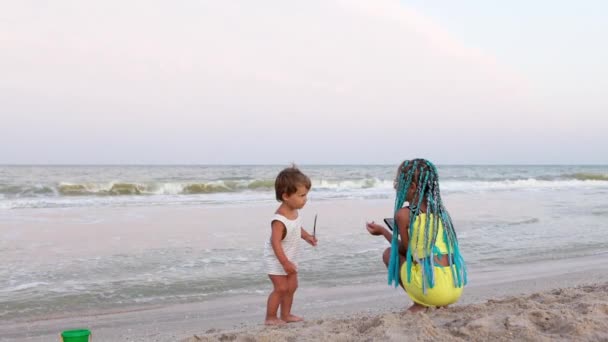 一个穿着白种人条纹T恤 一头金发的滑稽男孩与他的姐姐在海边的海滩上跳着欢快的现代音乐 她姐姐的辫子是非洲艳丽的 — 图库视频影像