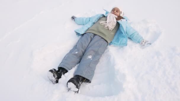 俯瞰着可爱的小女孩在新鲜的深雪中制造雪天使 穿着滑雪衣服和滑雪板的女孩躺着 挥动着胳膊和腿 在公园的雪地上成为天使 冬季活动的概念 — 图库视频影像