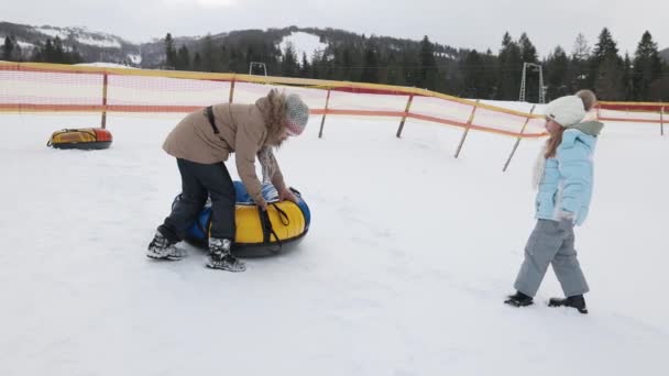 两个可爱的女孩在雪坡上提起黄色的雪管的侧视图 年龄较大的少女帮助她的妹妹继续前进 拉起滑雪板山 寒假的概念 — 图库视频影像