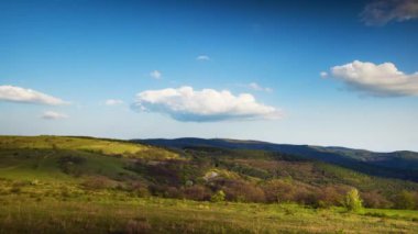 Valley with dry grassy green meadows with bare spring small bushes and big mighty colorful trees and low hilly Balkan mountains, under sky blue cloudy windy sky. 4K UHD timelapse video