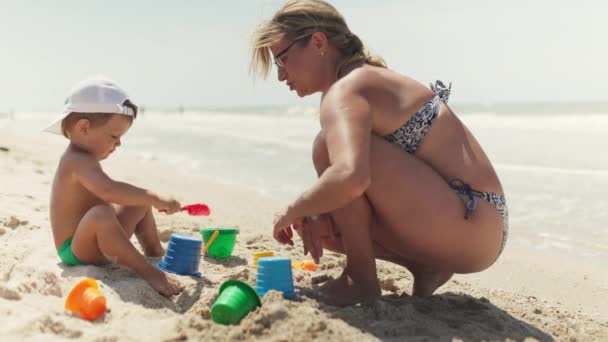 日焼けした幸せな思いやりのある母とともにブロンド髪で眼鏡で黒と青の水着は彼女の小さな面白い美しい赤ちゃんと遊んでいます 夏の海の近くの砂浜に座っています — ストック動画