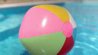 A cheerful rainbow bouncy ball floats, blown by the wind across a small childrens blue pool with clear clear water under the bright warm summer sun. HD slow-mo video