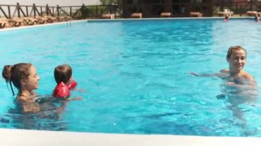 Happy caring kind mother in a bright bikini, plays and swims with a funny baby in oversleeves and his older sister in a deep blue pool with clear transparent water. HD slow-mo video