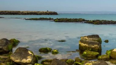 A wild stone and sandy beach with dark wet sea sand and large wet rocky stones near the reflective undulating spring Black Sea, under bright sunny sunset light in Bulgaria. 4K UHD timelapse video