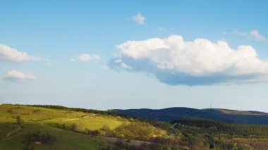 Valley with dry grassy green meadows with bare spring small bushes and big mighty colorful trees and low hilly Balkan mountains, under sky blue cloudy windy sky. 4K UHD timelapse video
