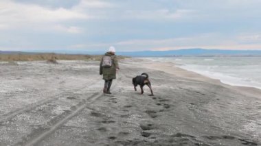 Ceketli enerjik neşeli genç kız mavi deniz kenarındaki kumlu sahilde sevgili sadık arkadaşı Rottweiler cinsinin büyük mutlu köpeği 4K UHD yavaş çekim videosu