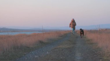 Ceketli enerjik neşeli genç kız mavi deniz kenarındaki kumlu sahilde sevgili sadık arkadaşı Rottweiler cinsinin büyük mutlu köpeği 4K UHD yavaş çekim videosu