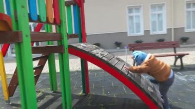 Cheerful active little boy in warm clothes plays on a bright playground in a kindergarten in an autumn empty park, and climbs a modern multi-colored slide in cloudy weather. 4K UHD realtime video