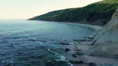 Rocky high sharp ledge with new fresh green vegetation over wild empty sandy beach in the shadow of a rock, near the deep calm undulating Black Sea under a clear blue bright sky. UHD 4K video realtime