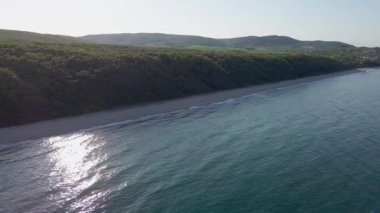 A wild empty sandy beach between a rocky sharp large ledge, covered with green fresh new vegetation, and a deep calm cool Black Sea under a clear blue sky. UHD 4K video realtime