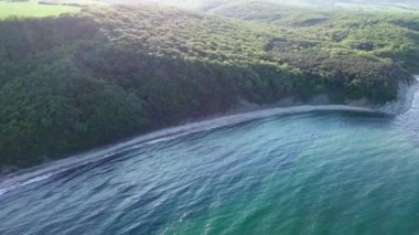 A wild empty sandy beach between a rocky sharp large ledge, covered with green fresh new vegetation, and a deep calm cool Black Sea under a clear blue sky. UHD 4K video realtime