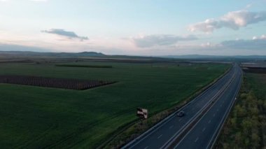 A smooth long road with modern powerful cars and trucks on it passes through farm industrial green fields, under an immense blue cloudy colored sky with a bright warm spring sun. UHD 4K video realtime