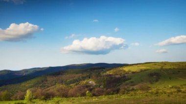 Valley with dry grassy green meadows with bare spring small bushes and big mighty colorful trees and low hilly Balkan mountains, under sky blue cloudy windy sky. 4K UHD timelapse video