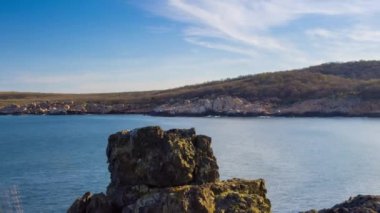 A wild stone and sandy beach with dark wet sea sand and large wet rocky stones near the reflective undulating spring Black Sea, under bright sunny sunset light in Bulgaria. 4K UHD timelapse video