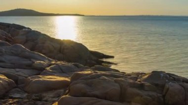 A wild stone and sandy beach with dark wet sea sand and large wet rocky stones near the reflective undulating spring Black Sea, under bright sunny sunset light in Bulgaria. 4K UHD timelapse video