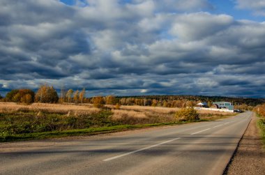 Yol, orman arazisi ve gökyüzü. Uzakta bir grup ev - sonbahar renklerinde bir manzara