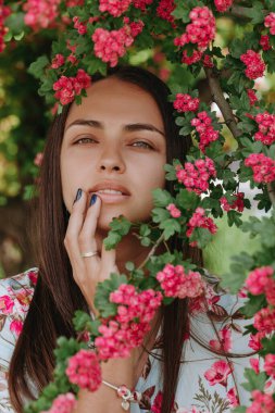 Güzel yeşil gözlü genç esmerin yakın portresi. Güzel Ukraynalı bir kadın dudaklarına dokunur ve yeşil çiçek bahçesinde poz verir. 