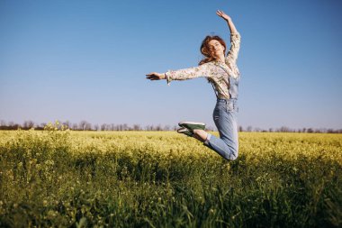 Neşe dolu genç bir kadın çiçek tarlasının ortasına atlıyor ve güneşin tadını çıkarıyor.