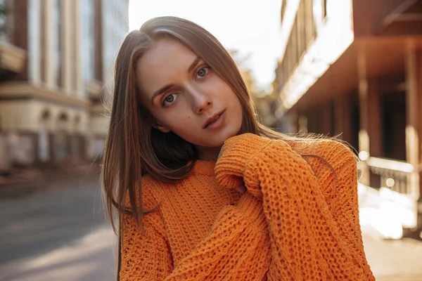 Genç bir kadın poz veriyor ve kameraya bakıyor. Turuncu uzun kollu kazak giymiş bir manken. 