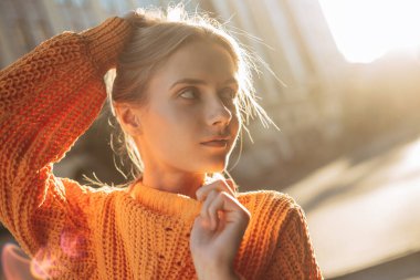 Kız bir eliyle başını, diğer eliyle yakasını tutuyor ve hafifçe gülümsüyor. Turuncu, sıcak süveter giyen genç kadın modelin yakın portresi