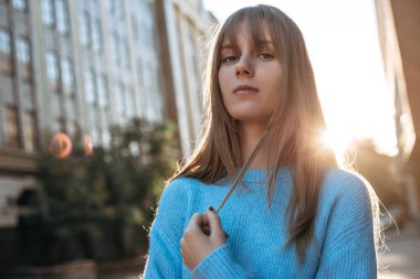 Uzun sarı saçlı, kendine güvenen genç bir kadın kameraya bakıyor ve şehirde poz veriyor. Saç kesimi püsküllü ve mavi kazaklı bir kadın model. 