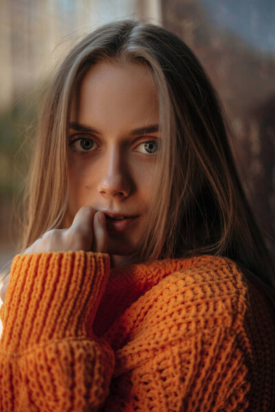 Милая и романтичная молодая женщина с длинными светлыми волосами, одетая в теплый оранжевый вязаный свитер с длинным рукавом, смотрящая в камеру с загадочным видом