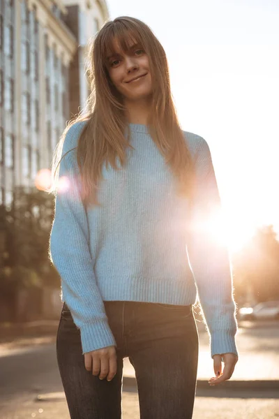 正の若い女性ですオーバーサイズブルーセーター立って屋外で街の夕日を背景にカメラを見て笑顔 ストックフォト
