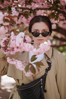 Siyah güneş gözlüklü romantik bir kadın. Bel çantalı. Çiçekli dallarda dikilip kameraya bakıyor.