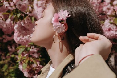 Young pretty woman with flower behind the ear posing in the blooming cherry garden. Female model head profile shot