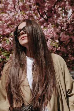 Sakura ağaçlarının parkında güzel bir kadın model poz veriyor. Mantolu ve güneş gözlüklü modacı kadın..