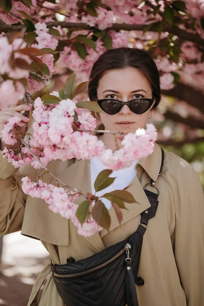 Siyah güneş gözlüklü romantik bir kadın. Bel çantalı. Çiçekli dallarda dikilip kameraya bakıyor.