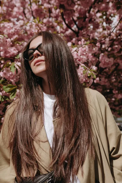 Sakura ağaçlarının parkında güzel bir kadın model poz veriyor. Mantolu ve güneş gözlüklü modacı kadın..