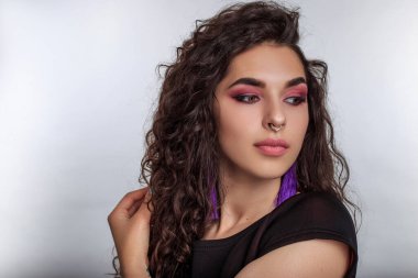 Uzun siyah kıvırcık saçlı, profesyonel makyajlı ve beyaz arka planda stüdyoda poz veren şehvetli bir kadın model..