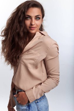 Bluzlu, kot pantolonlu, fotoğraf stüdyosunda beyaz arka planda duran ve kameraya bakan kendine güvenen genç bir kadın. 