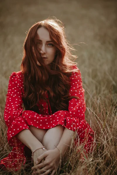 美しい若い大人の女性の赤いドレス小麦の分野で彼女の足の上に交差腕とカメラを見て座って — ストック写真