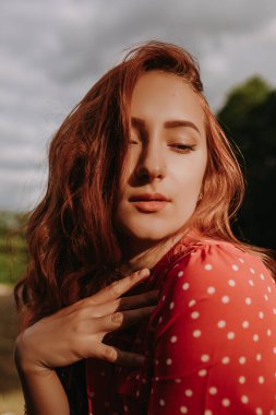 Kızıl saçlı kadın, ormanın arka planındaki kuru çim tarlasında kamera önünde poz veriyor.