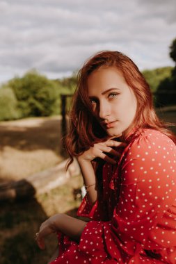 Kırmızı elbiseli, beyaz noktalı, kırmızı saçlı, kurumuş ağaçta oturan ve kameraya poz veren genç ve güzel bir kadın. Kadın model yüzüne dokunuyor ve kameraya bakıyor.