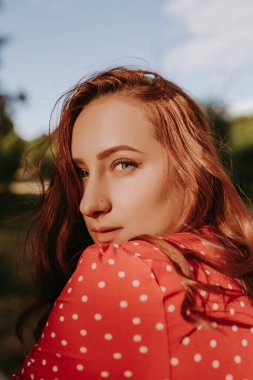 Kızıl saçlı kadın omzunun üzerinden kameraya bakıyor. Uzun kıvırcık saçlı, kırmızı elbiseli, beyaz noktalı bir kadın.