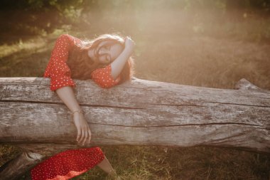 Genç ve güzel bir kadın, kırmızı yaz elbisesi ve beyaz benekleri ile ormanın ortasında kurumuş bir ağaç gövdesinde yatıyor.