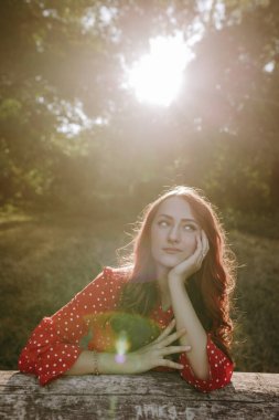 Kızıl saçlı, güzel bir kadının portresi. Kırmızı tişörtlü, tahta kirişe yaslanmış ve kameraya poz veriyor.