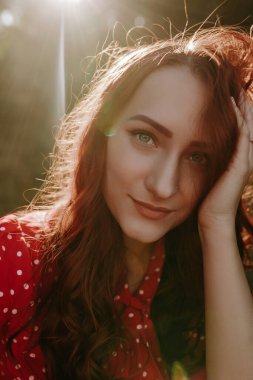 Kapalı yüz portresi güzel beyaz beyaz kadın kırmızı uzun kıvırcık saçlı, kırmızı gömlekli, başını tutuyor ve kameraya bakıyor.